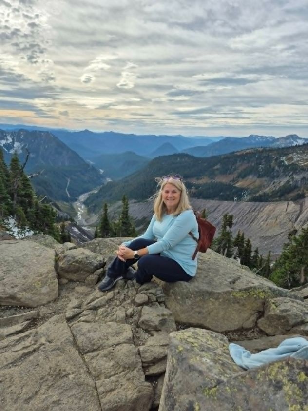 Joy smiling on a rocky overlook at Mount Rainier with mountains and a river valley behind her