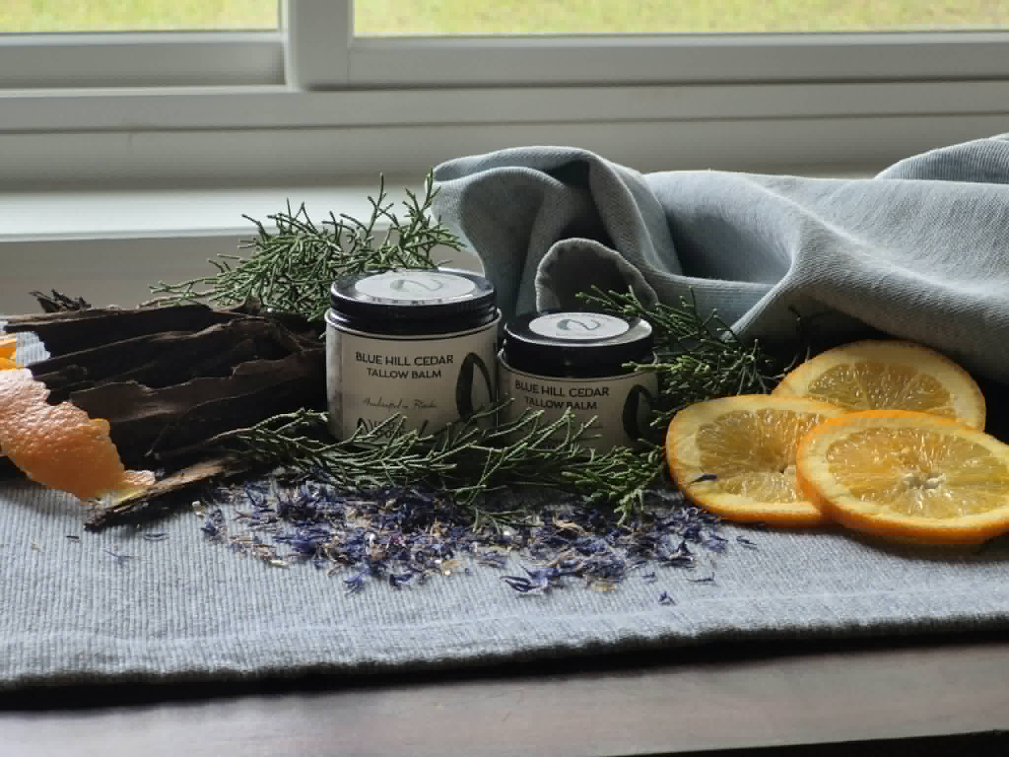 Two jars of Blue Hill Cedar tallow balm with cedar sprigs, bark, cornflower petals and orange slices